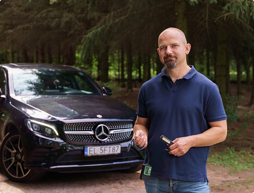 Marek Ejsmont stoi przy ciemnym Mercedesie GLC w lesie, trzymając kluczyki do auta